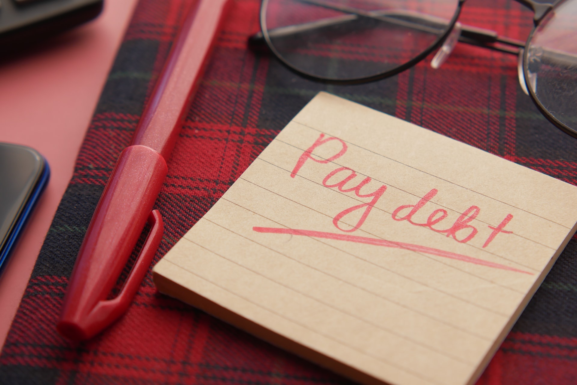 Pay debt written on sticky note in red pen on top of red plaid notebook